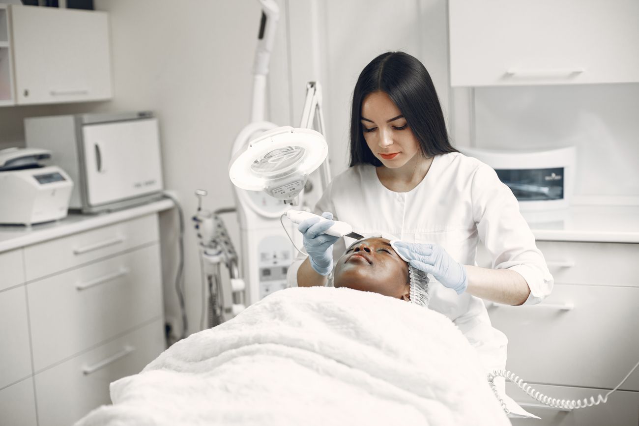 An aesthetician in a white coat giving a client a facial treatment in a bright, modern treatment room